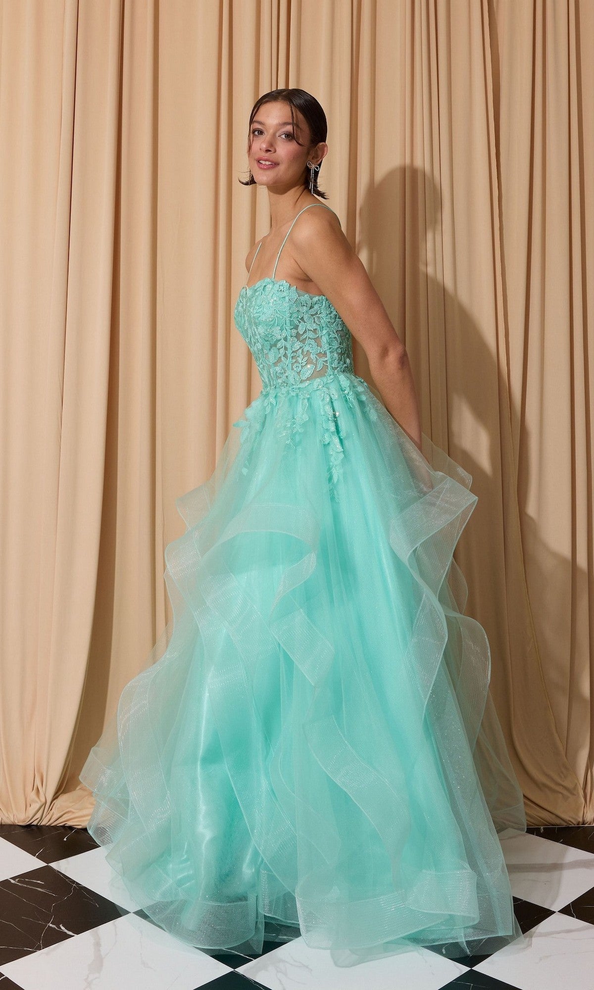 A woman poses in the Eureka EK167 Long Prom Dress, a light blue strapless gown with a floral lace embroidered corset and ruffled tulle skirt, in front of beige curtains on a black and white tiled floor.
